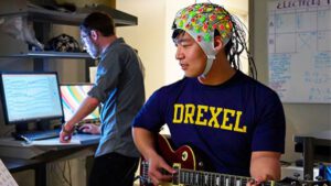a man with tan skin and black hair wearing an EEG cap with colorful electrodes on it as he plays a guitar. A researcher stands in the background at a computer, watching brain waves on the screen