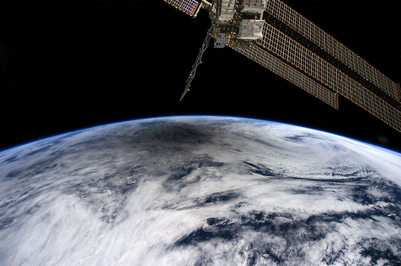 The space station's solar array wings can be seen in the foreground. in the background clouds on Earth with the shadow of the moon on them