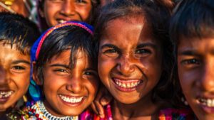 Close-up image of a group of four Indian children (with the face of another more blurred behind them) who are smiling at the camera. The sun in shining on their faces